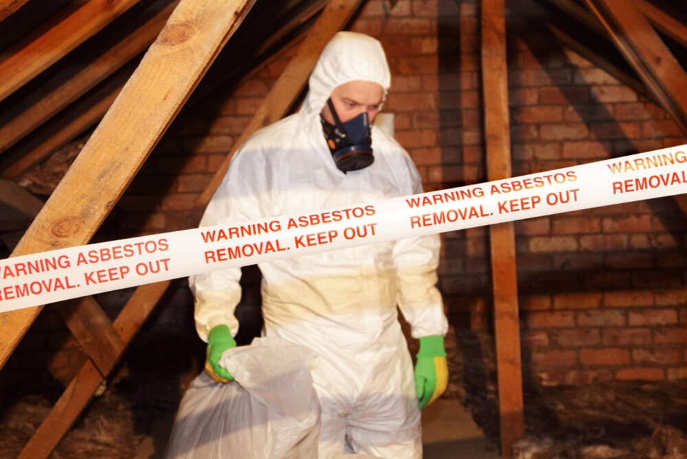 A worker in full protective gear while performing asbestos removal. The area is sectioned off with red and white warning tape that reads “WARNING ASBESTOS REMOVAL. KEEP OUT.”