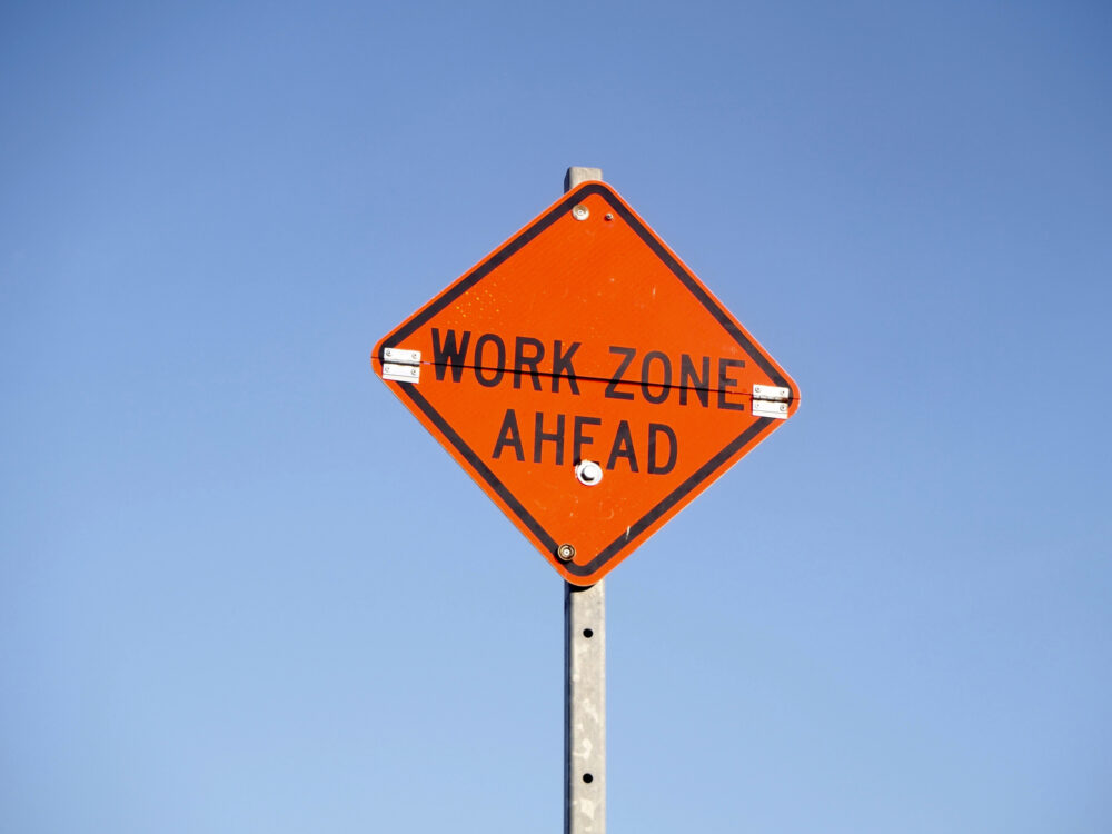 An orange, diamond-shaped sign with the words “WORK ZONE AHEAD”.