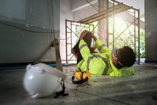 A worker in reflective clothing has fallen from scaffolding and is on their back on the ground holding their leg in pain.