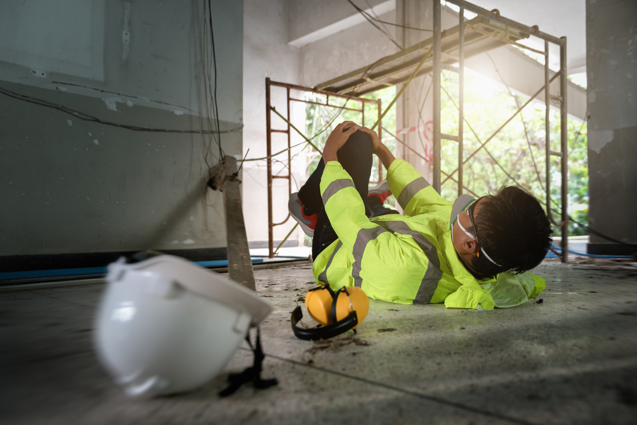A worker in reflective clothing has fallen from scaffolding and is on their back on the ground holding their leg in pain.