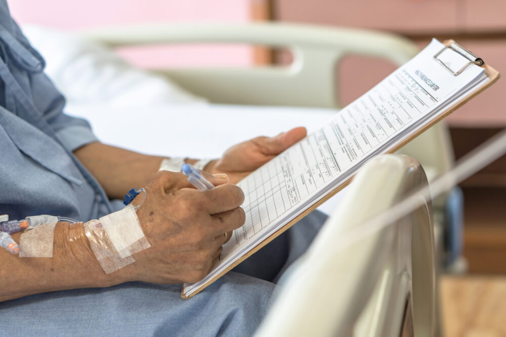 An elderly patient connected to an IV drip is in a hospital bed, filling out paperwork on a clipboard.