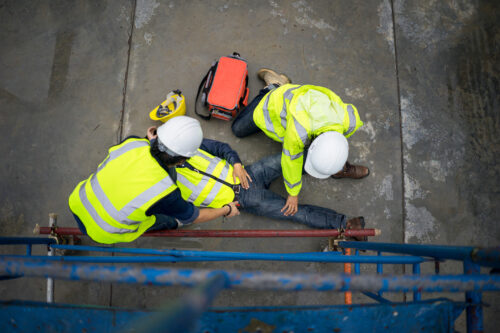 Two construction workers in hard hats and reflective vests examine a fallen, presumably injured, coworker. There is a red duffel bag next to them.