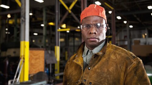 A Massachusetts factory worker wearing a hard hat and protective eyewear facing the camera with the factory equipment in the background.