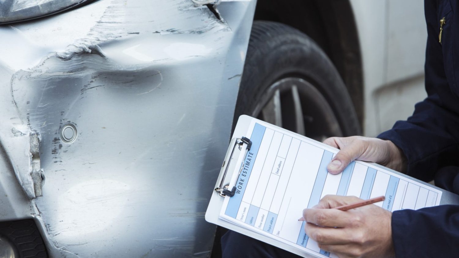 A claims adjuster inspecting damage to a vehicle, holding a clipboard and writing on a paper titled, “WORK ESTIMATE.”