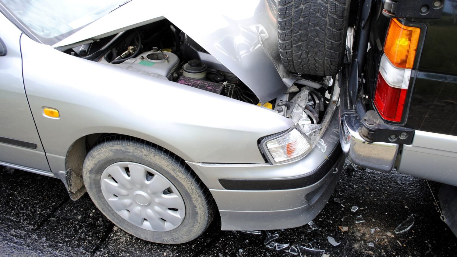 A silver car with a damaged front bumper after colliding with the rear of a black SUV in a rear-end accident.