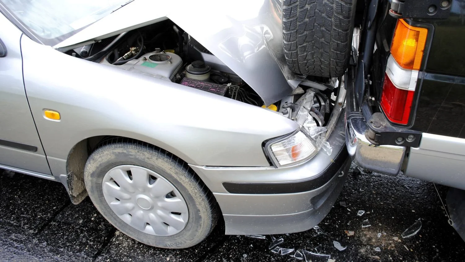 A silver car with a damaged front bumper after colliding with the rear of a black SUV in a rear-end accident.
