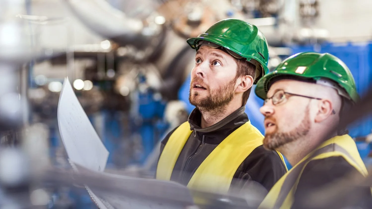 Two construction workers with hard hats hold papers and evaluate working conditions.