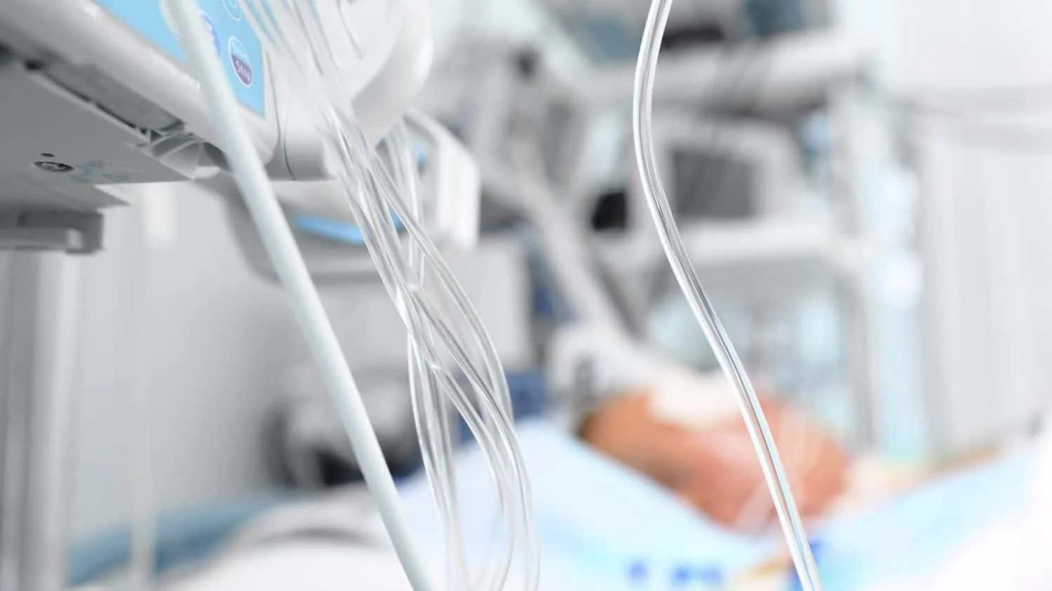 Tubes attached to a hospital bed with a patient in the background.