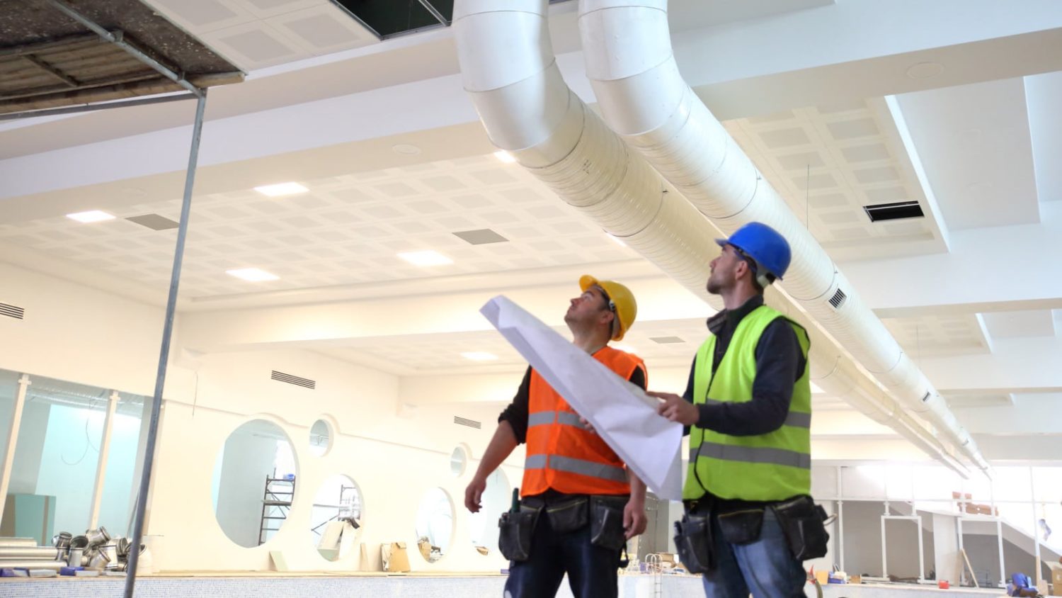 Two construction workers in reflective vests look up at the ceiling, with one holding blueprints.