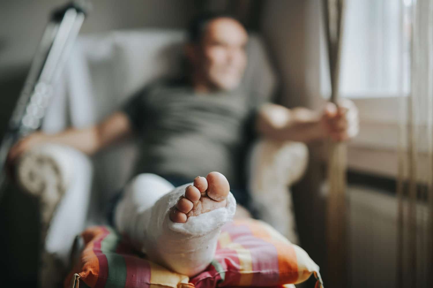 A man sits in a chair with his leg elevated and wrapped in a cast, crutches leaning against the chair. The focus is on the injured leg in the foreground.