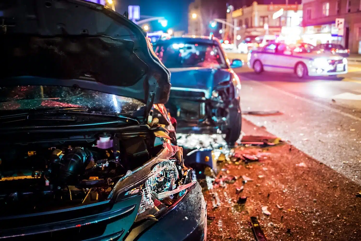 Two badly damaged vehicles on the side of the road in a city at nighttime. A police car can be seen to the right.