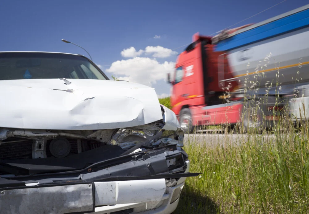 The aftermath of a Cambridge, MA, truck accident which has left a white car badly damaged. A commercial truck is seen passing in the background.