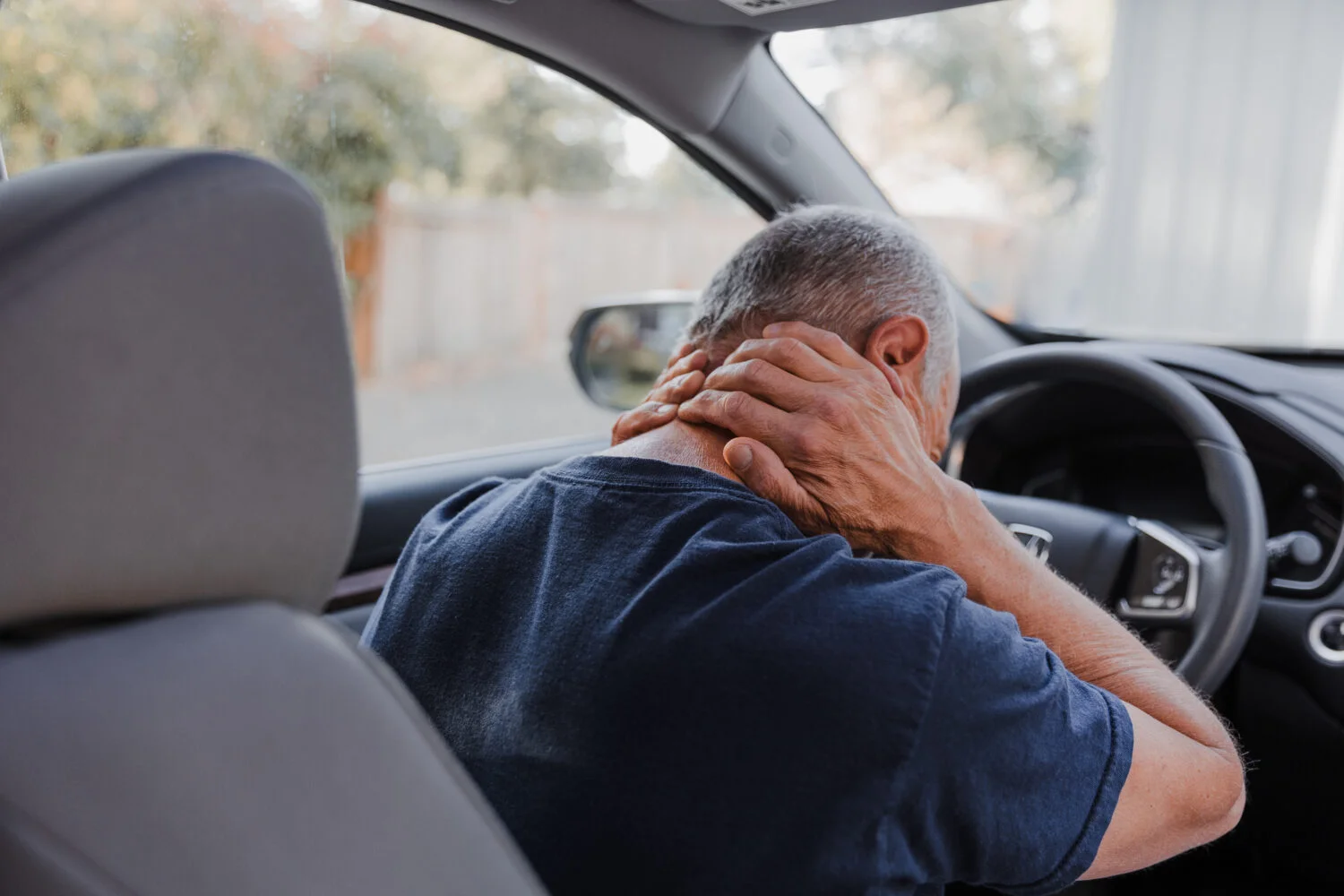 A man sits in the driver seat of his vehicle while holding the back of his neck in pain.