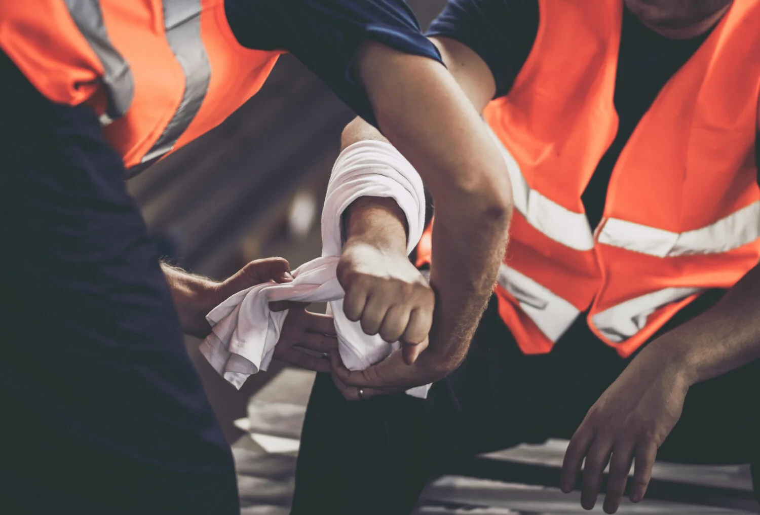 Two workers in reflective vests. One is bandaging the arm of the other who has been injured.