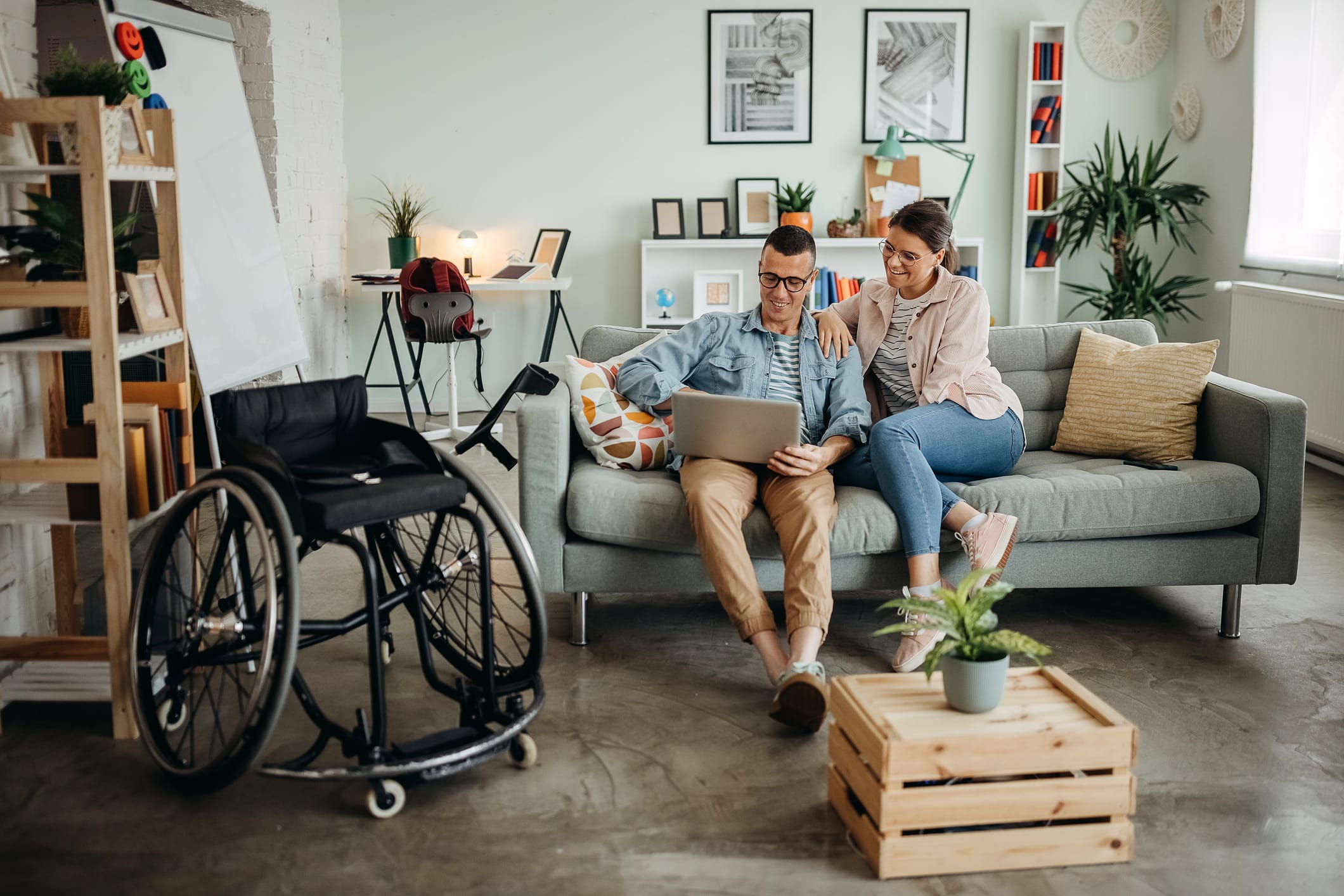 A man applies for disability on his laptop while he and a woman are sitting on the couch. A wheelchair is seen next to them.