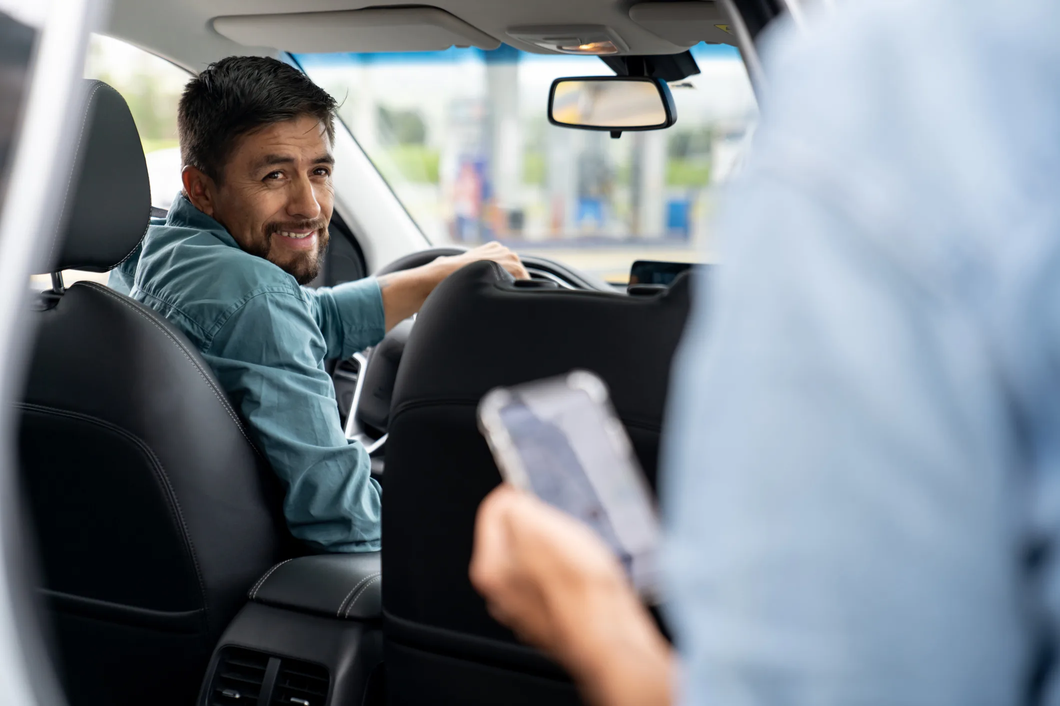 A smiling rideshare driver turns around to face their passenger who is looking at their phone.