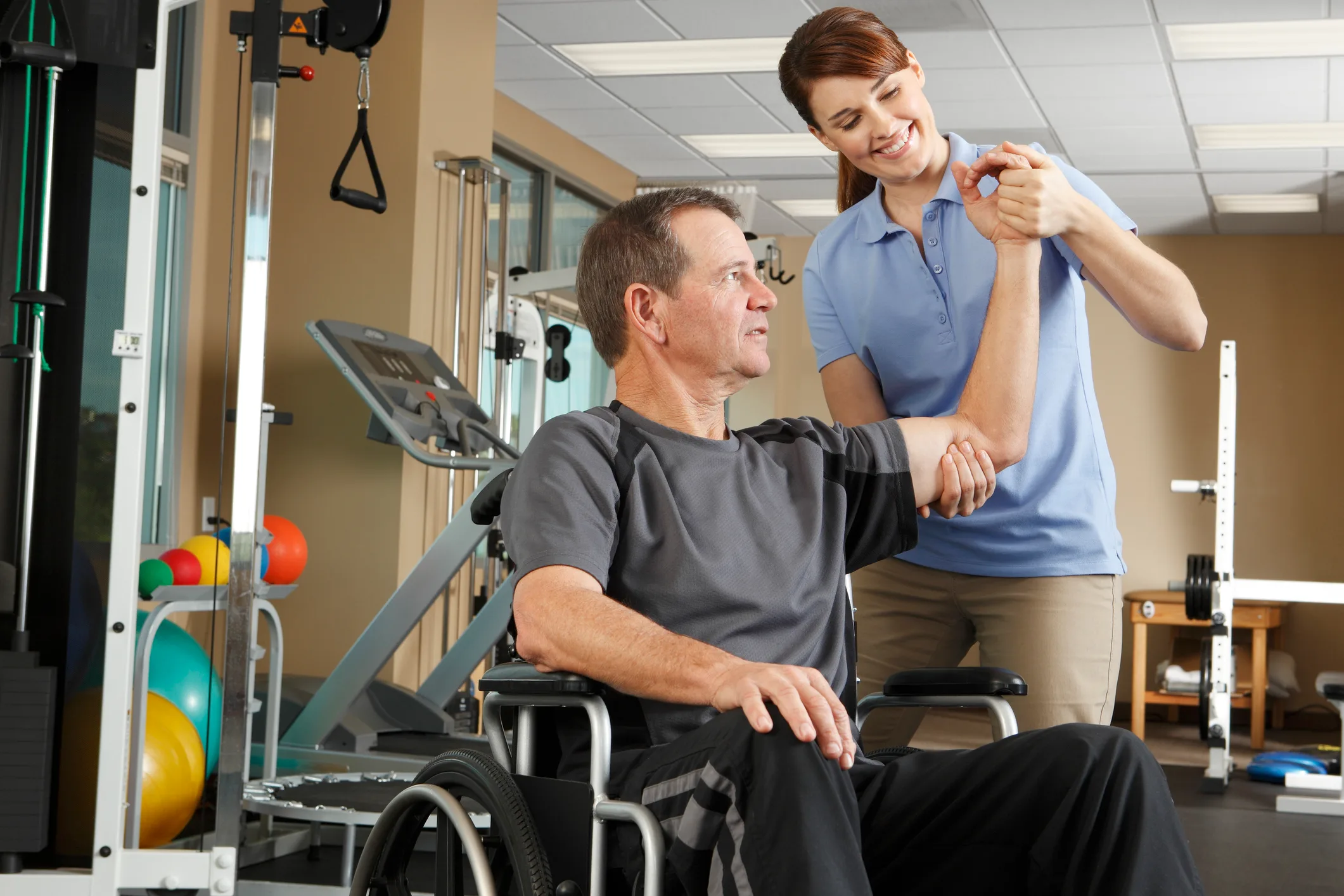 A physical therapist holding a man’s hand during a session. The man is in a wheelchair and the two are in a rehabilitation facility.