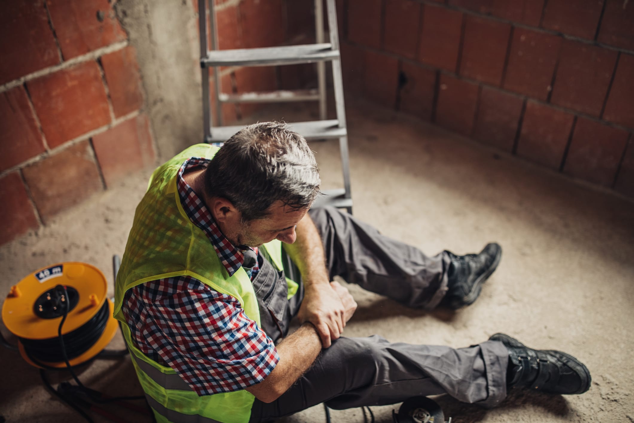 A construction worker sits on the ground in visible pain, holding his injured arm while wearing a safety vest. The ladder nearby indicates a recent fall.