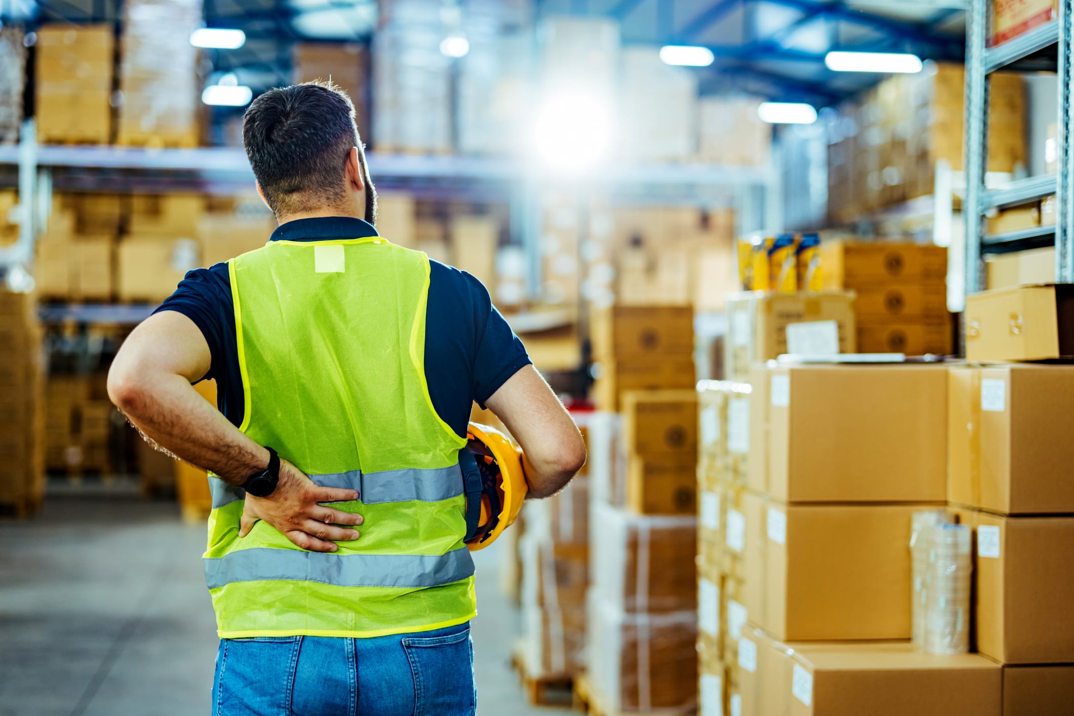 A warehouse worker in a reflective vest, holding his back with one hand (as if injured) and holding a helmet with the other. Boxes are stacked all around.