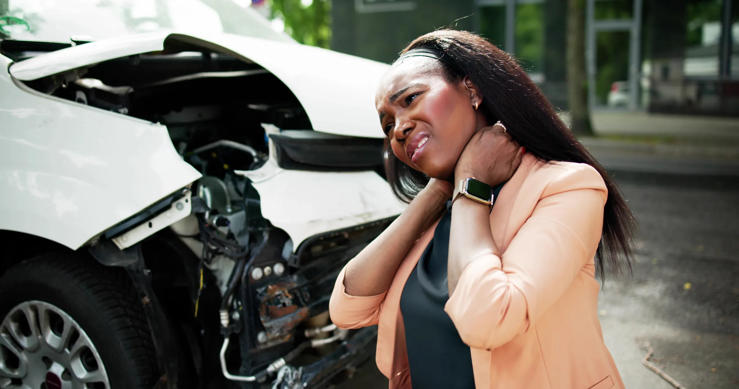 A woman holds her neck in pain in front of a badly damaged car, indicating a recent wreck.