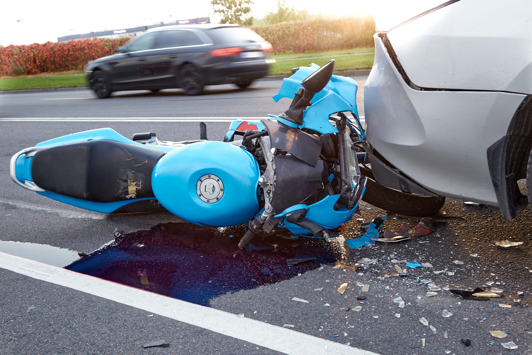 A badly damaged blue motorcycle lies on the ground under the bumper of a car. Another vehicle can be seen driving on the road in the background.