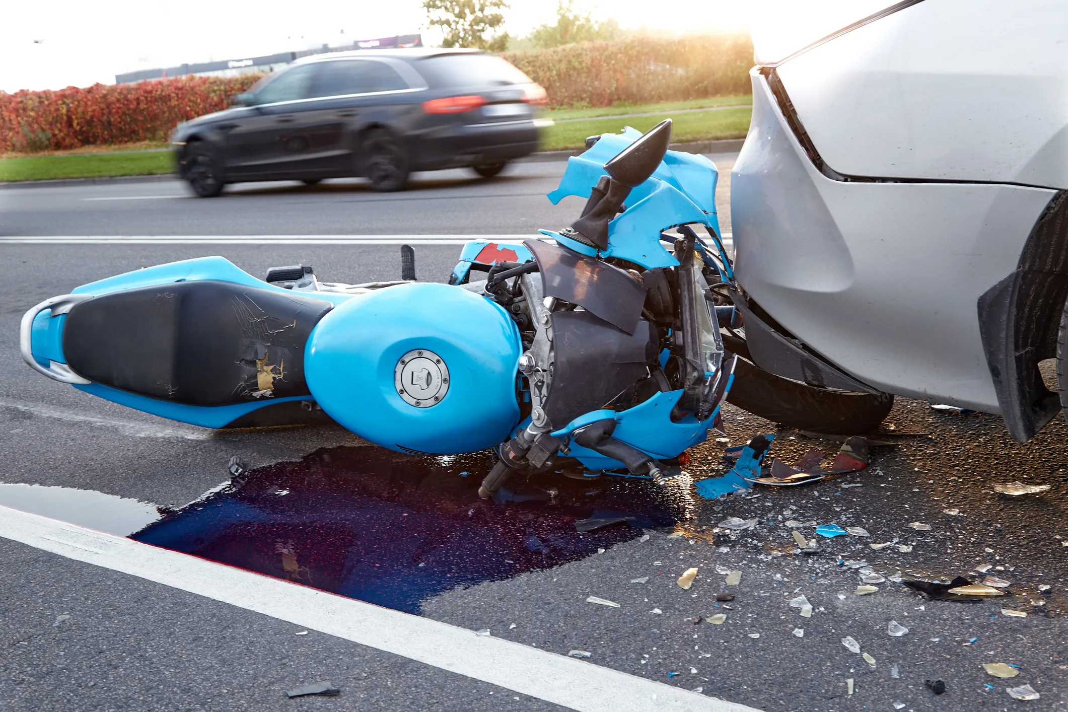 A badly damaged blue motorcycle lies on the ground under the bumper of a car. Another vehicle can be seen driving on the road in the background.