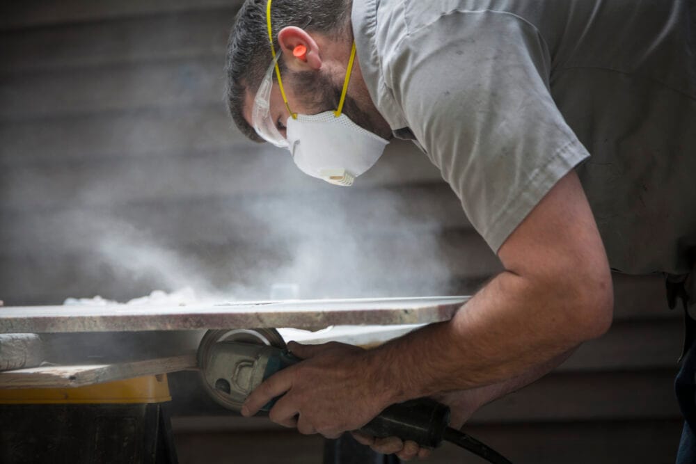 A cloud of dust rises as a worker wearing a mask and safety glasses uses a grinder to cut through a stone slab.