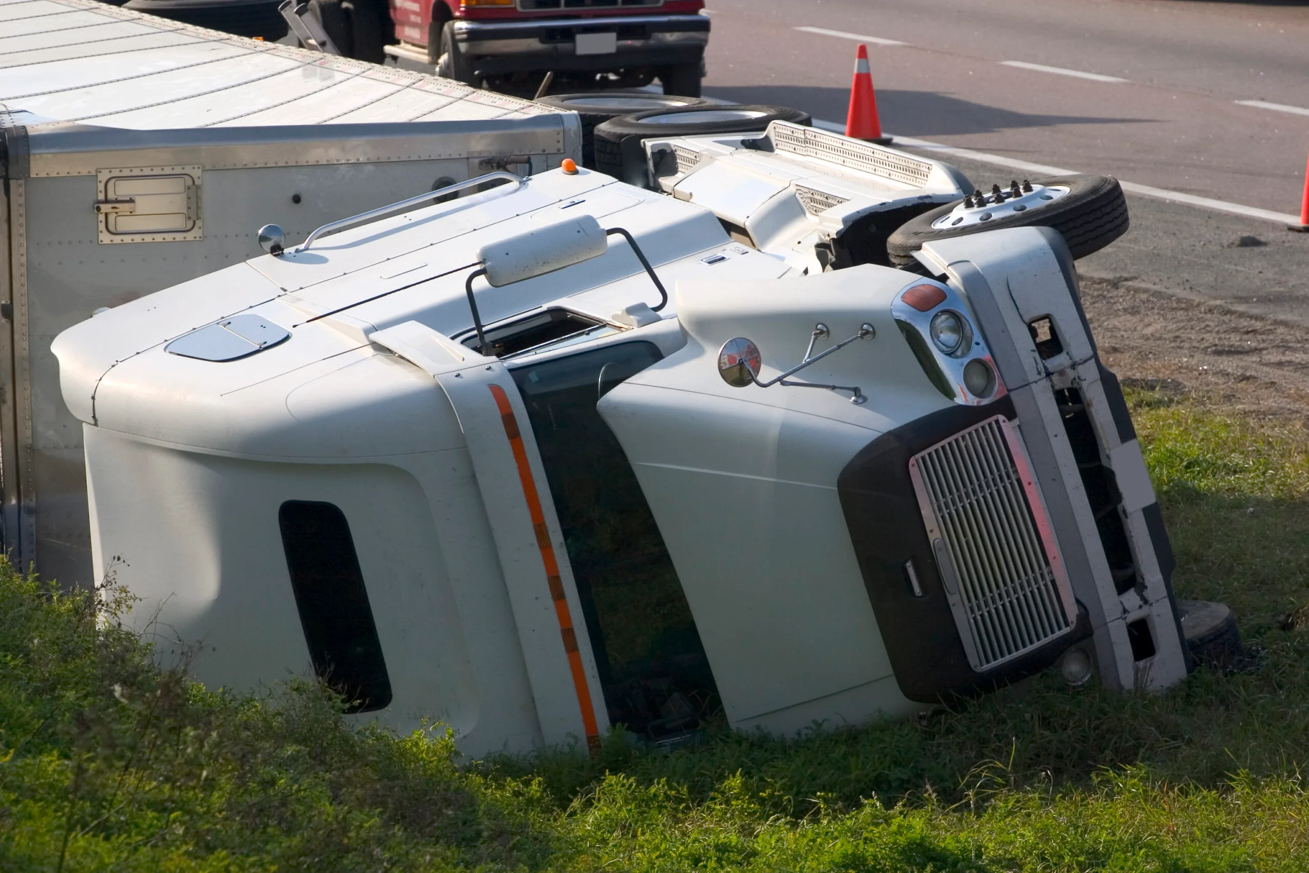 An overturned white tractor-trailer on the side of the road. An emergency vehicle and orange cones can be seen in the background.