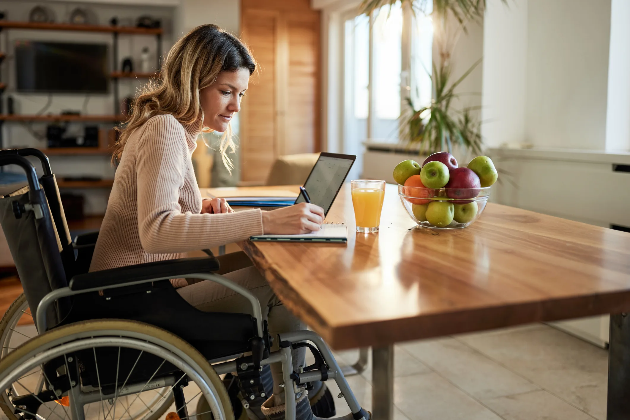 Woman in a wheelchair sitting at a kitchen table, writing in a notebook while using a tablet, with a glass of juice and a bowl of fruit nearby.