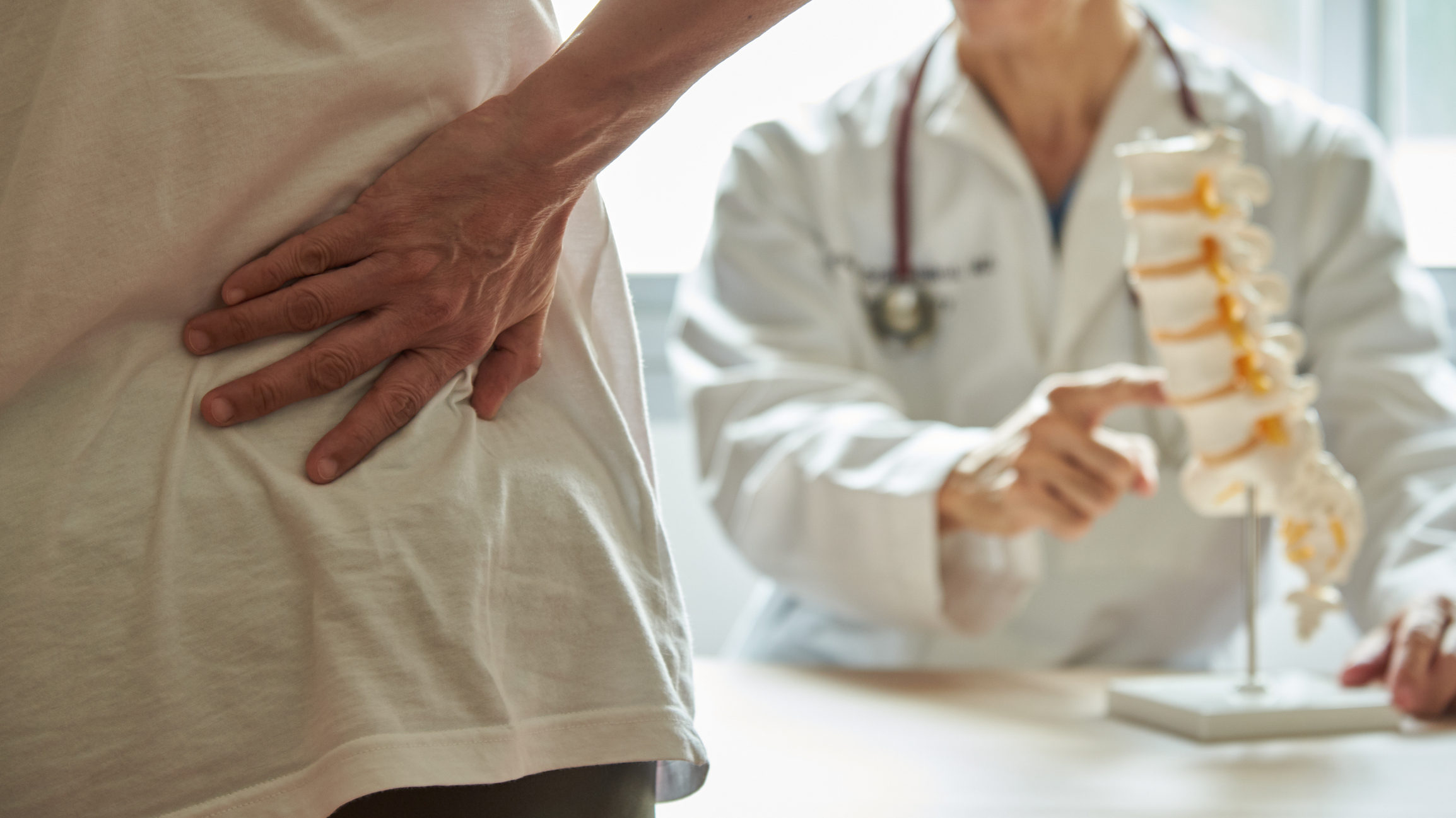 A person holding their lower back in pain stands in front of a doctor who is pointing to a model of the spine during a medical consultation.
