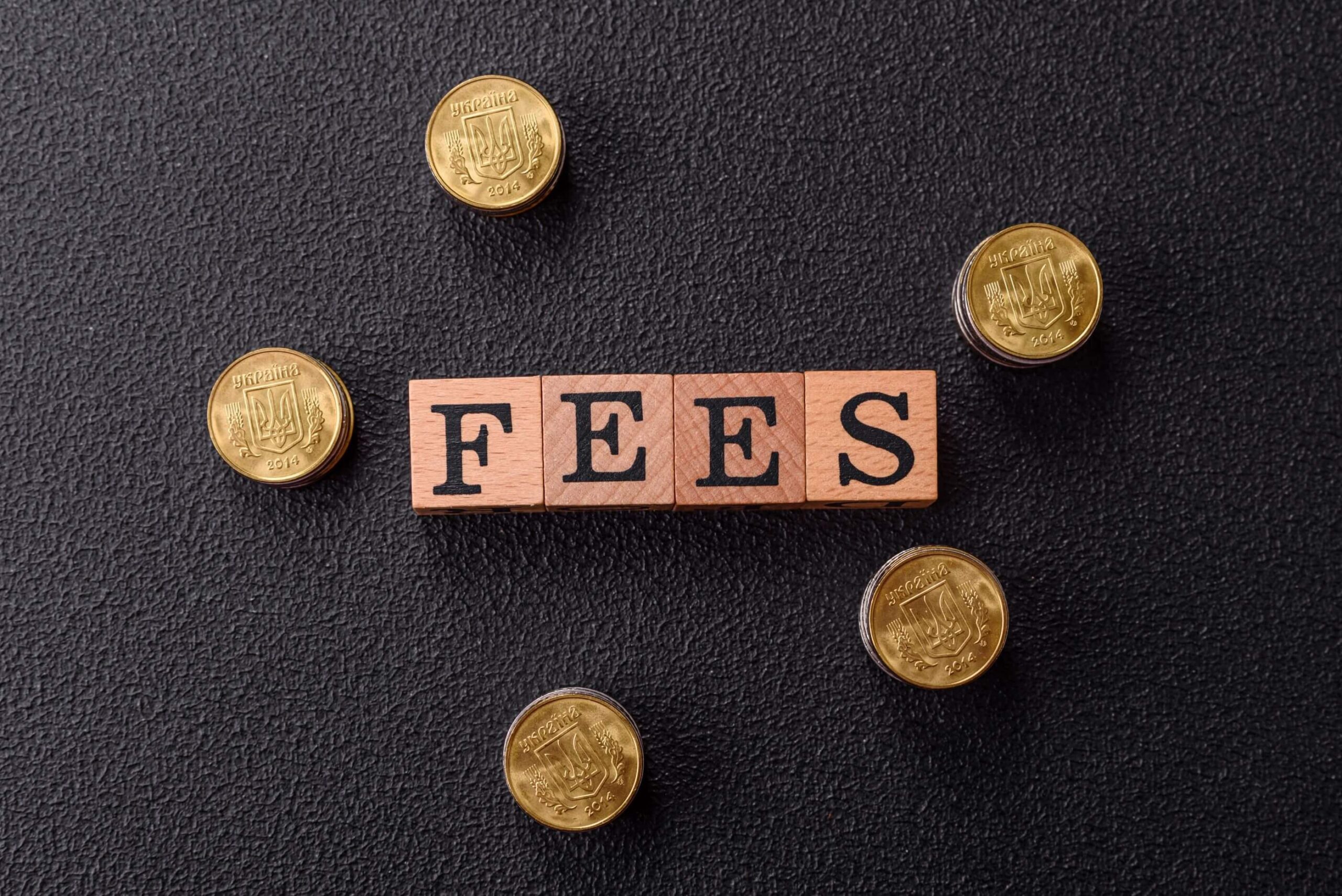 Wooden blocks surrounded by stacks of gold coins spell out the word “fees.”