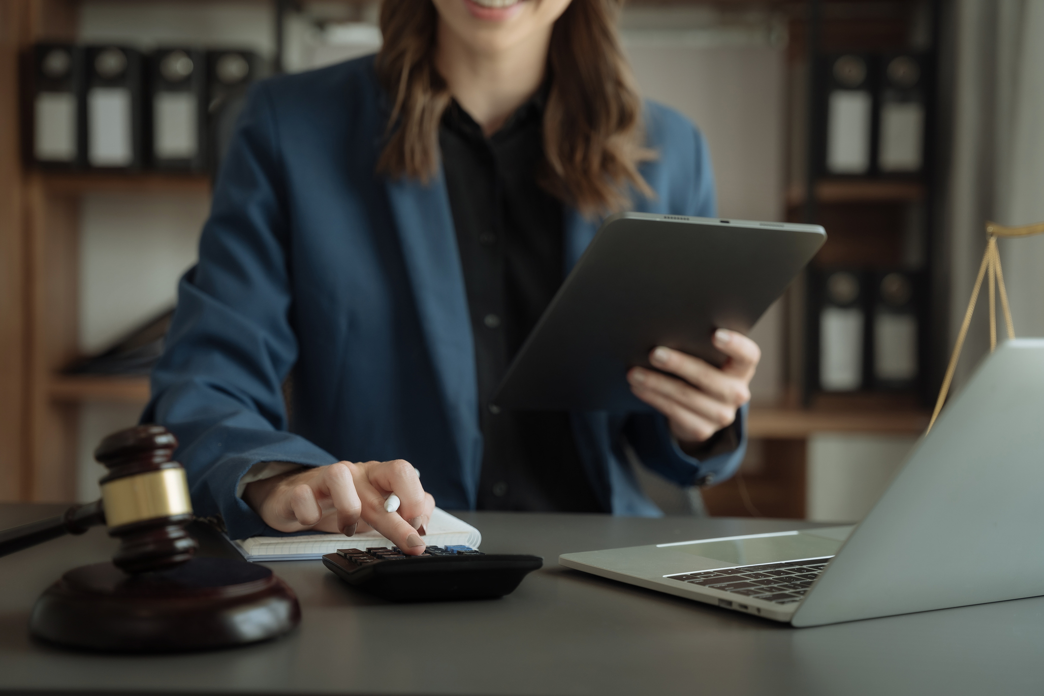 A lawyer calculating the possible settlement amount for a client. She’s typing on a calculator while holding a tablet. A gavel, a notebook, and a laptop are sitting on her desk.