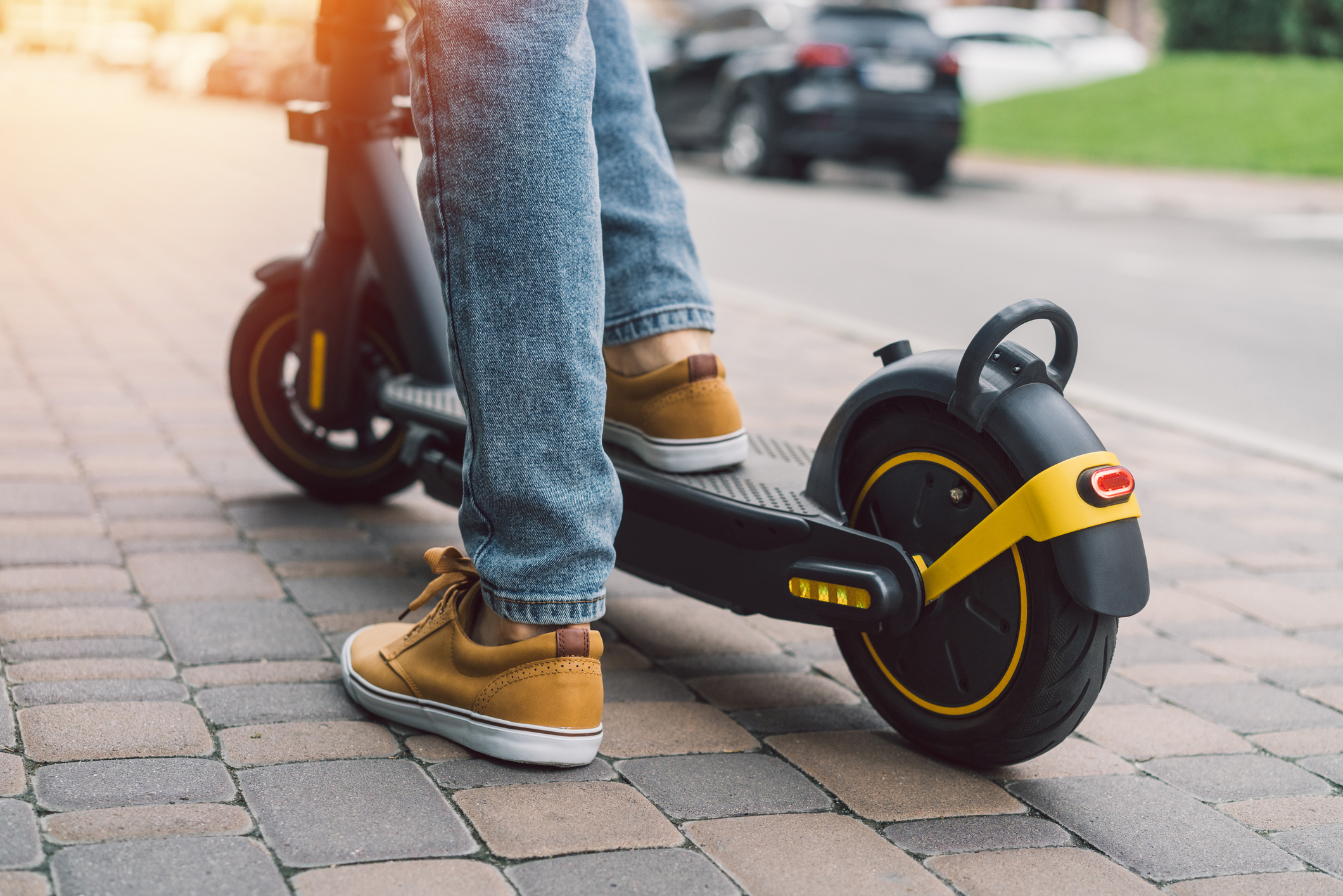 Knee-down view of a person riding an electric scooter down a paved path. Cars can be seen in the background.