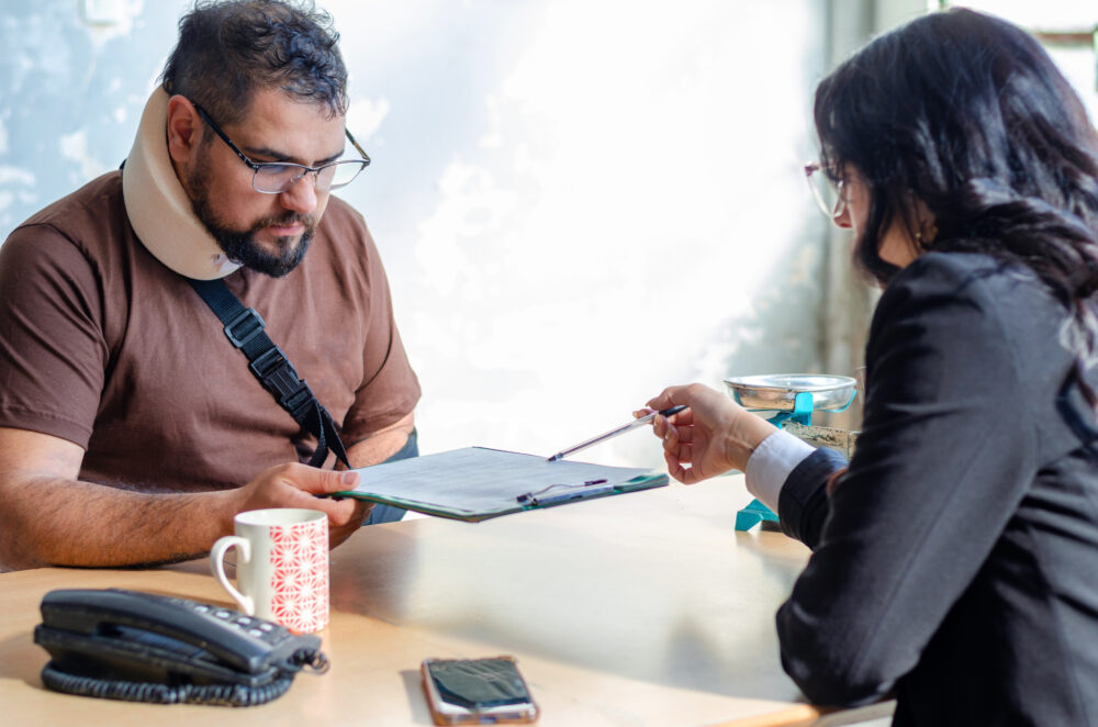 A car accident lawyer and her injured client review paperwork on a clipboard. A landline, a cellphone, and a mug are on the desk next to them.