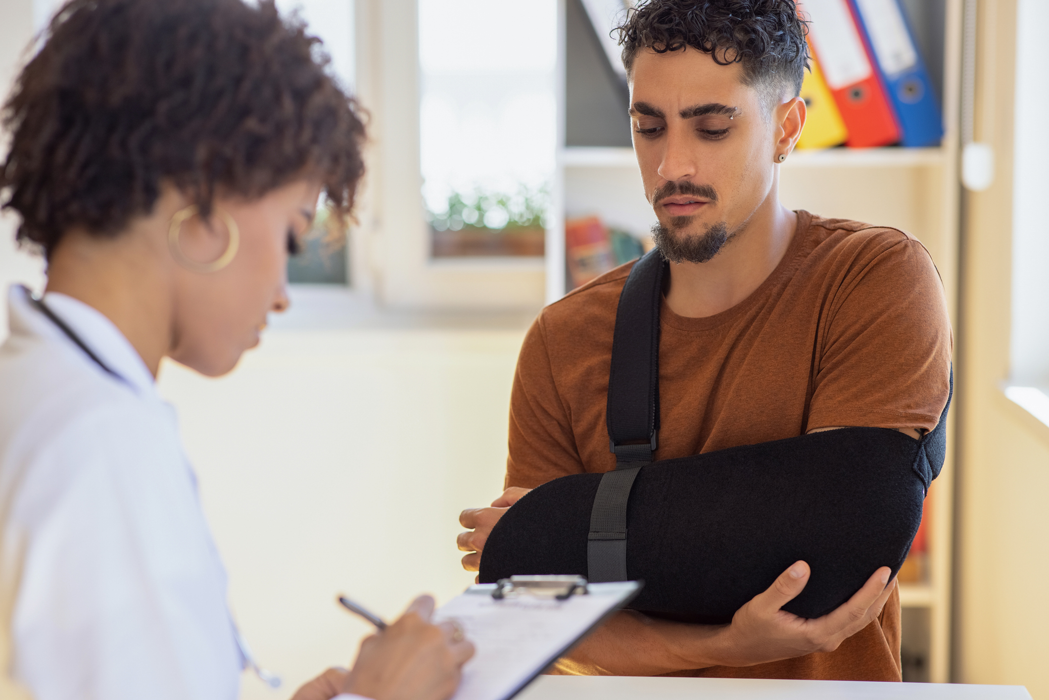 A man with his arm in a sling after a work injury is being evaluated by a physician with a clipboard.