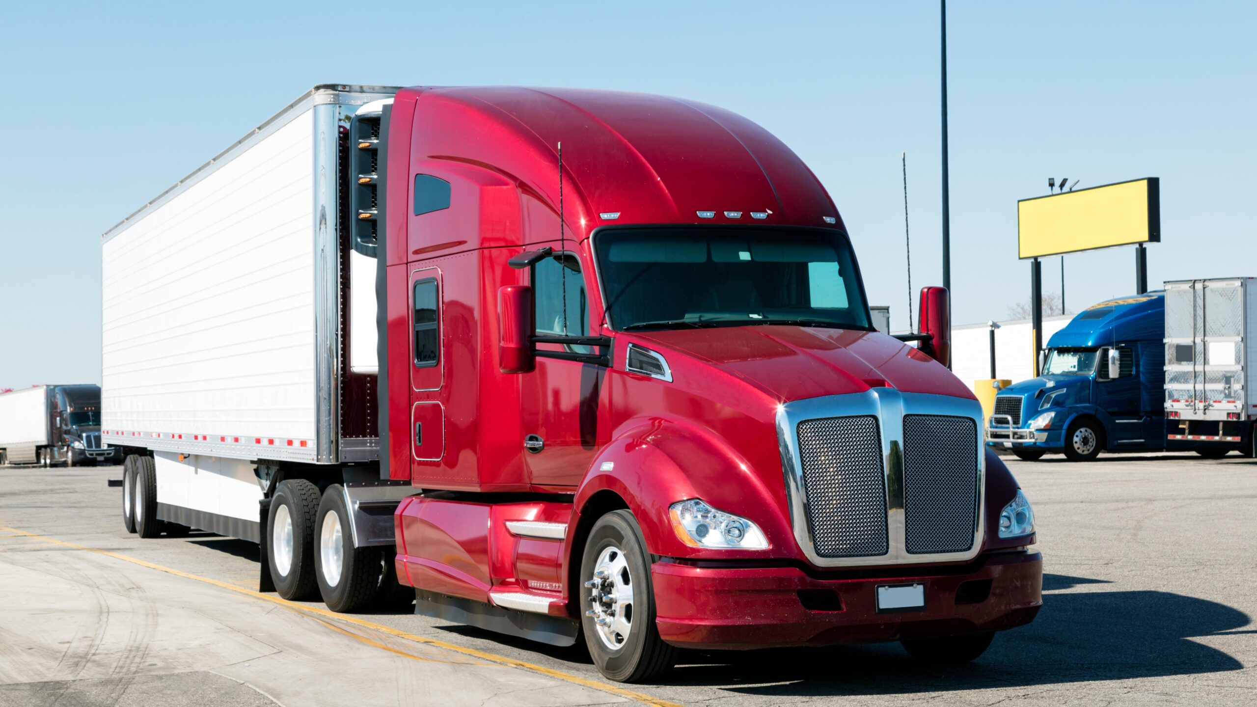 A red big rig with a white trailer. In the background, other commercial trucks can be seen.