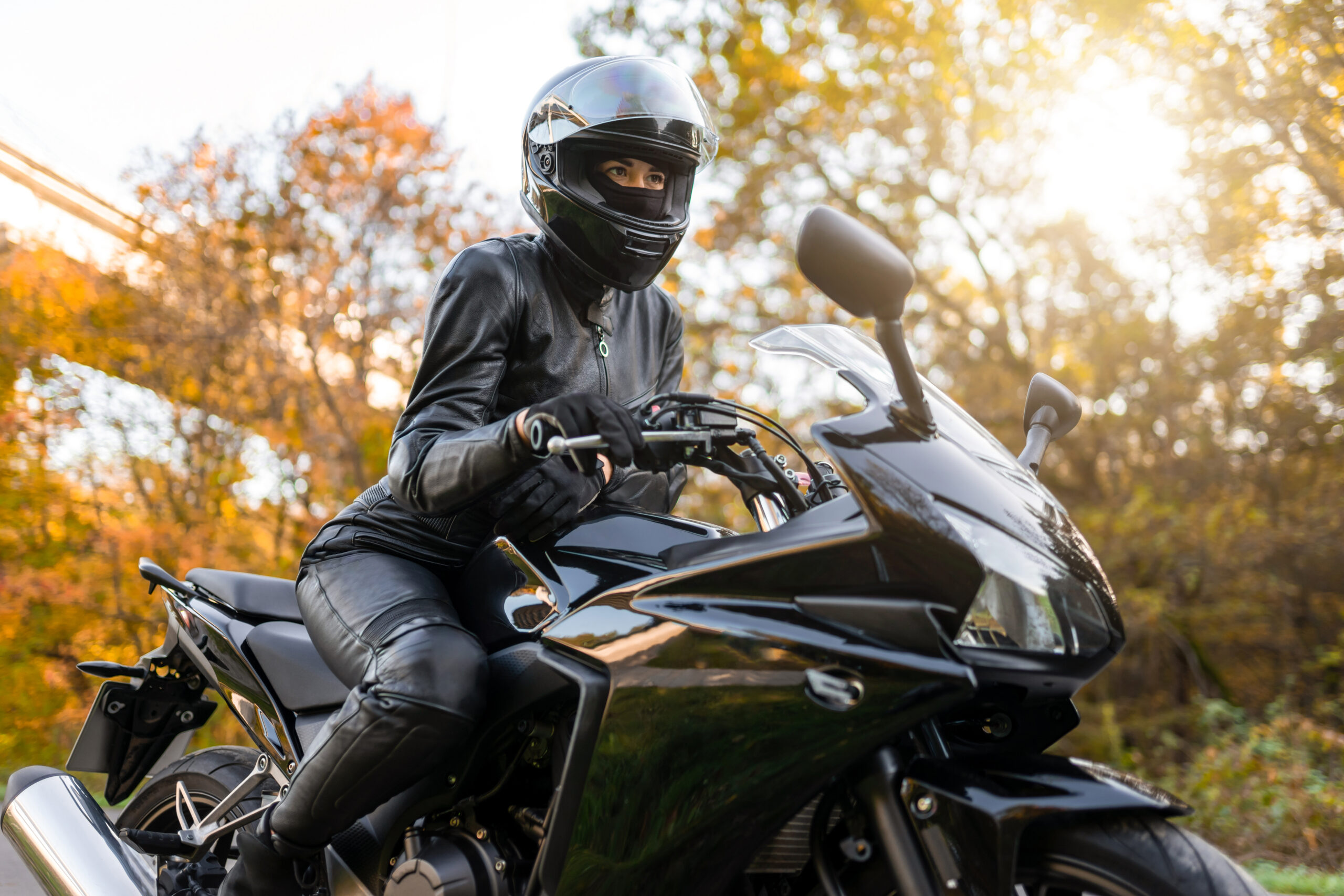 A motorcycle rider in all black, on a black motorcycle with trees and a bridge in the background.