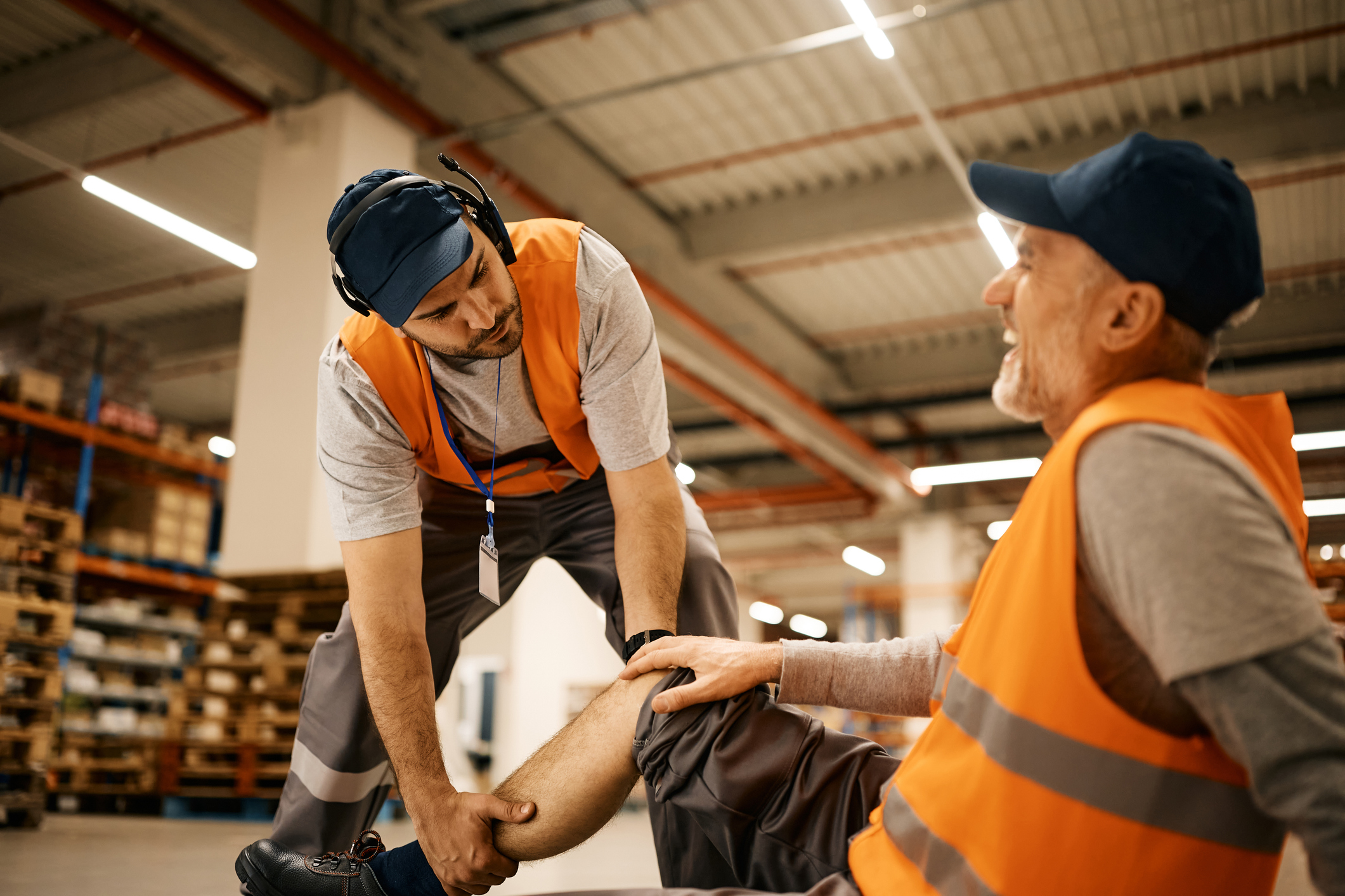 Two warehouse workers in orange vests, one has fallen and is holding his knee, the other is stabilizing the fallen man’s leg.