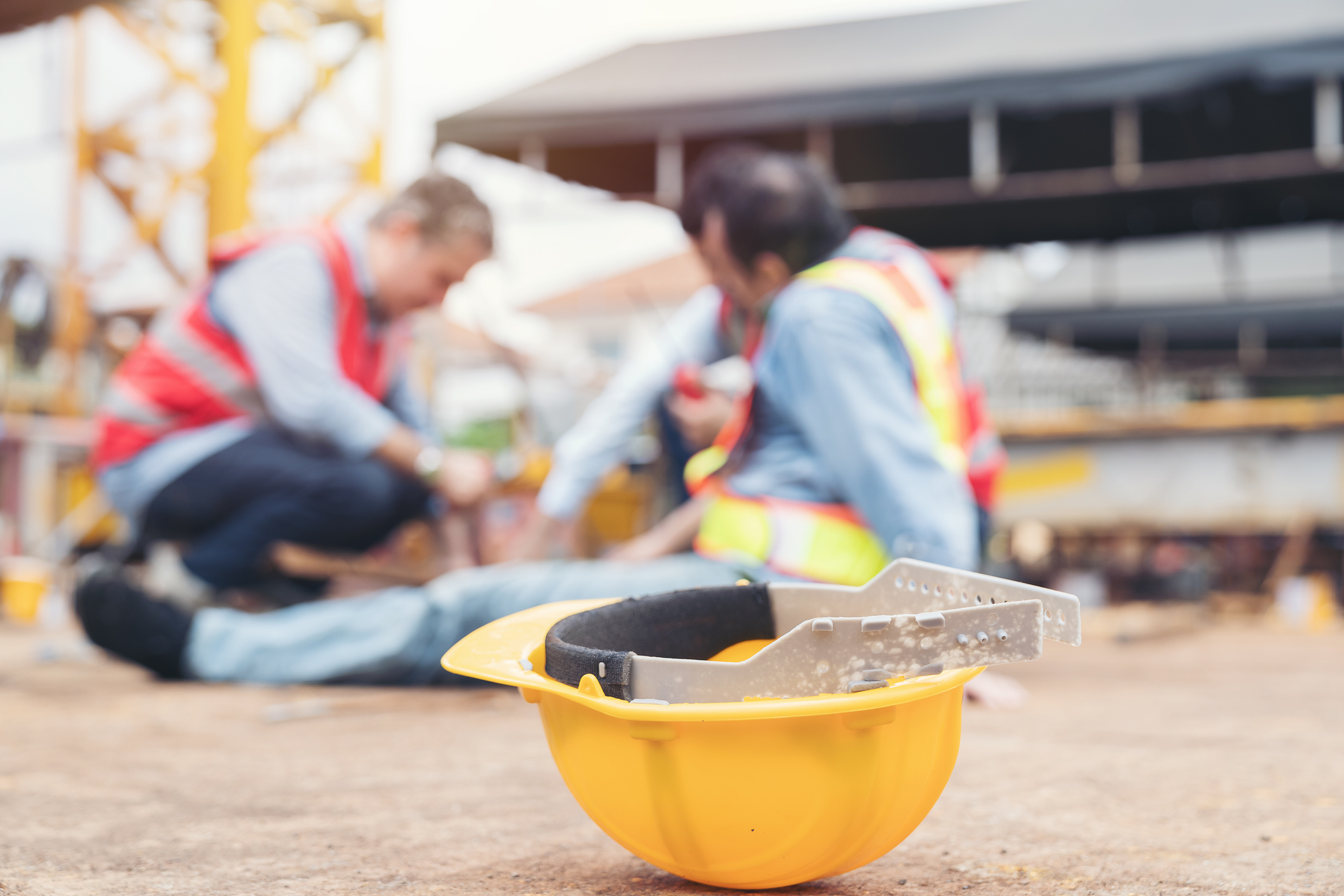 A hard hat is in the foreground, while a fallen worker and his coworker tend to the fallen worker’s injuries in the background.