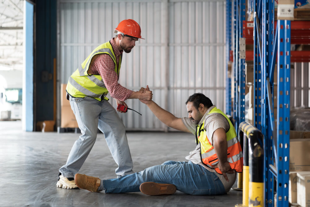 A worker with a hard hat and walkie-talkie helps his coworker up after a fall.