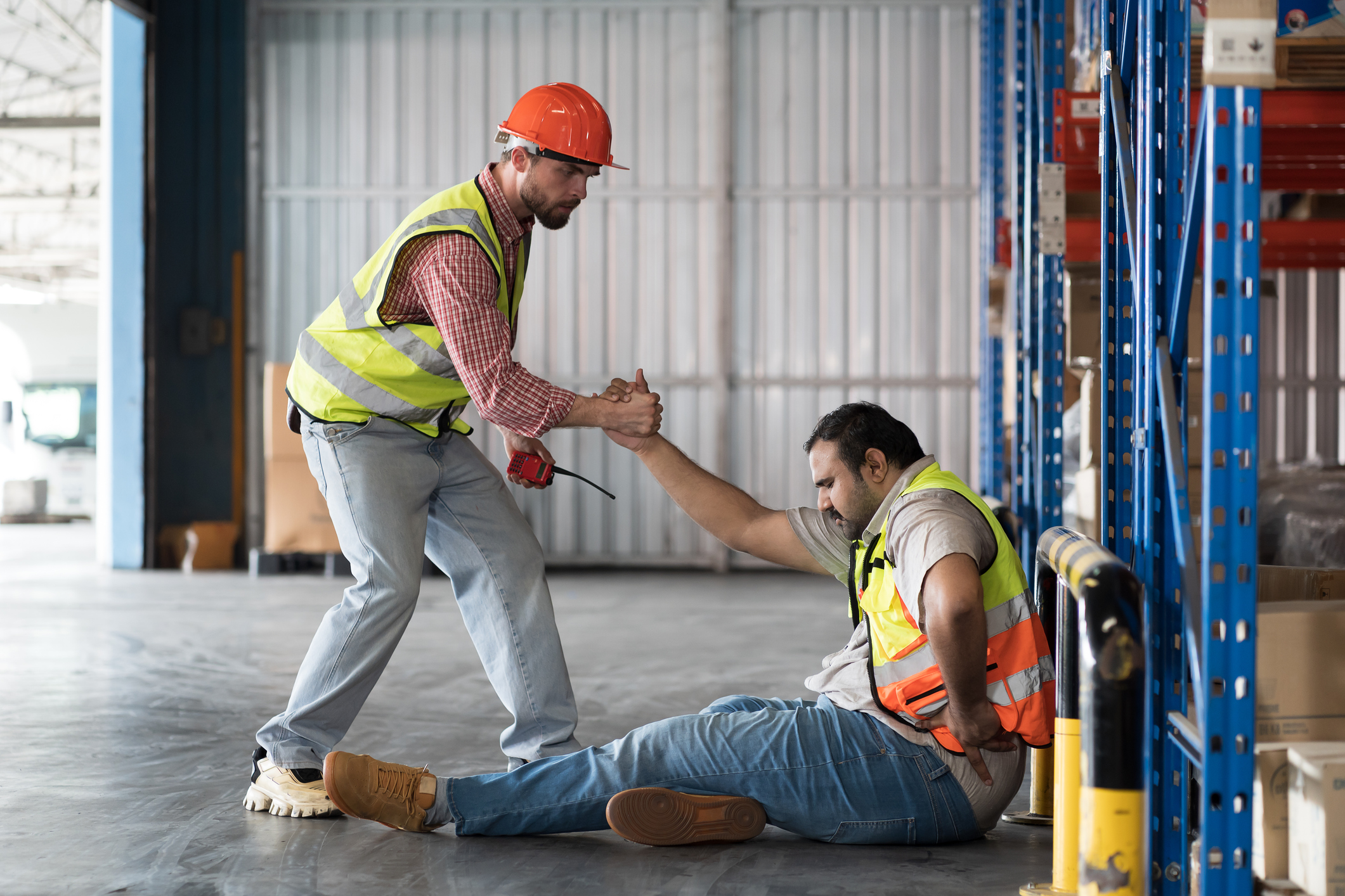 A worker with a hard hat and walkie-talkie helps his coworker up after a fall.
