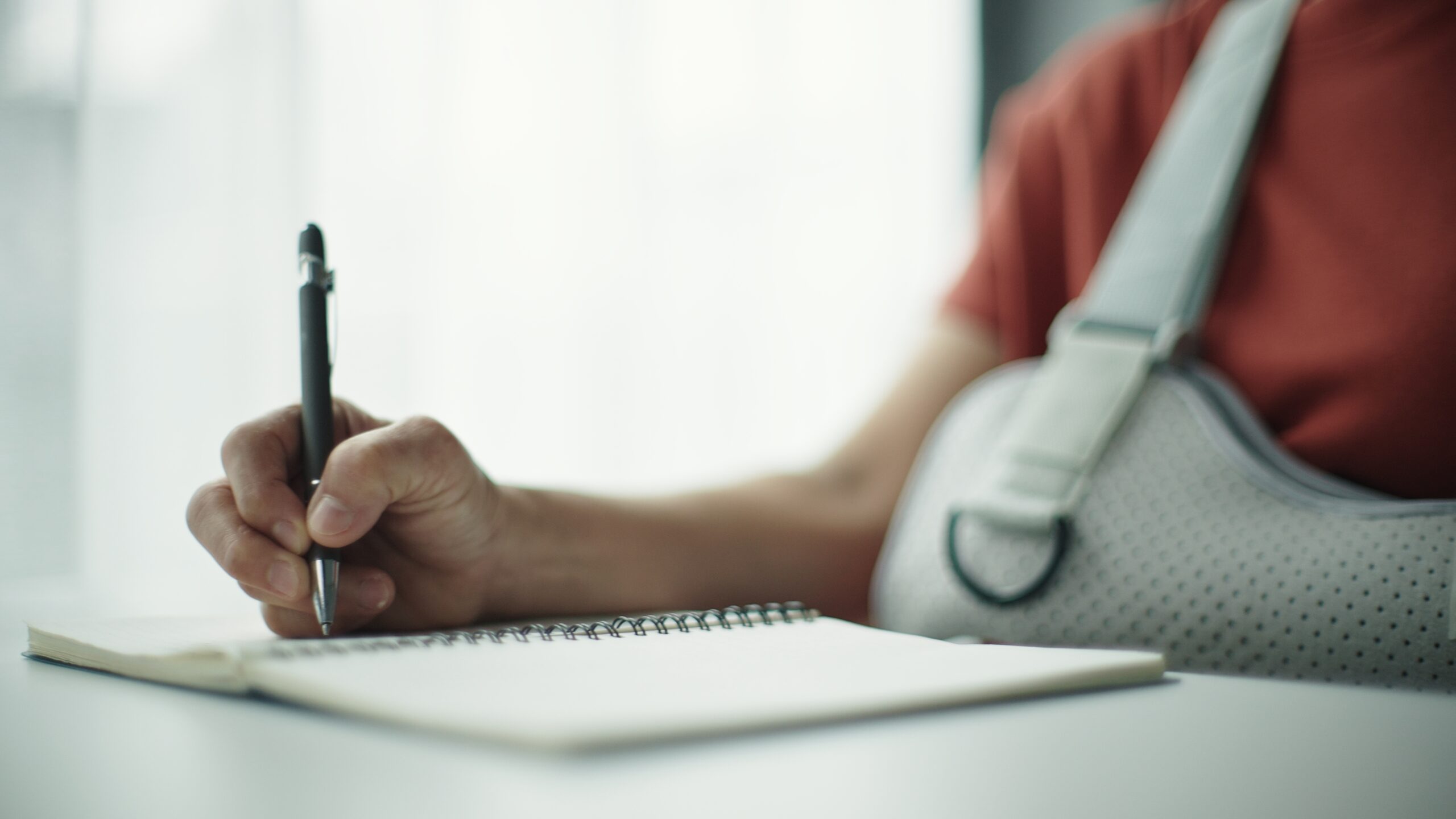 An individual with their arm in a sling writes in a notebook.