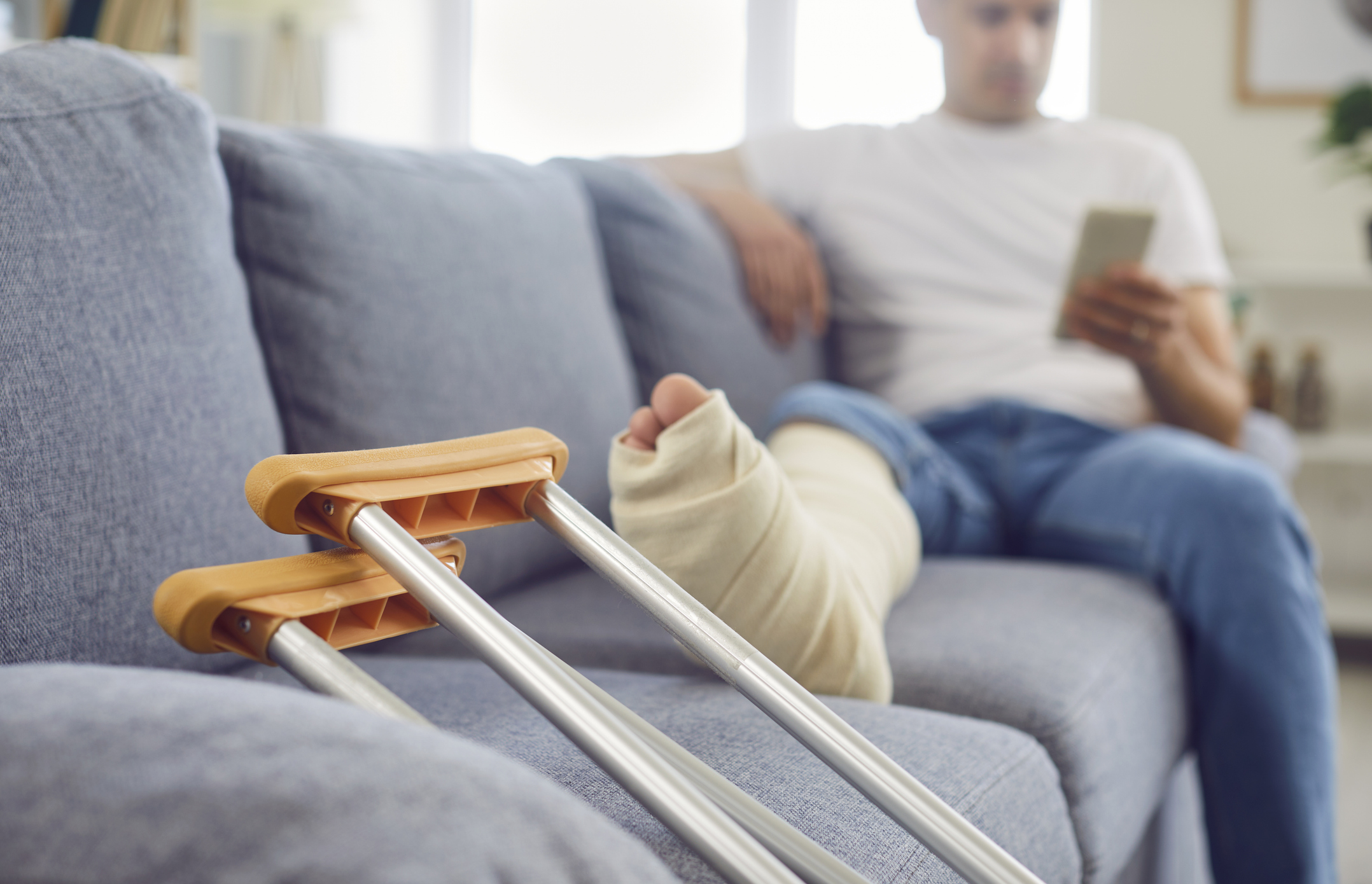 An individual with a bandaged leg sits on the couch, looking at their phone. Crutches are leaned against the couch.