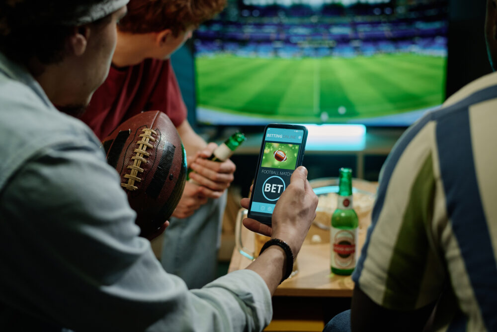 Friends watching football with snacks and beers, while one places a bet through a mobile betting app.