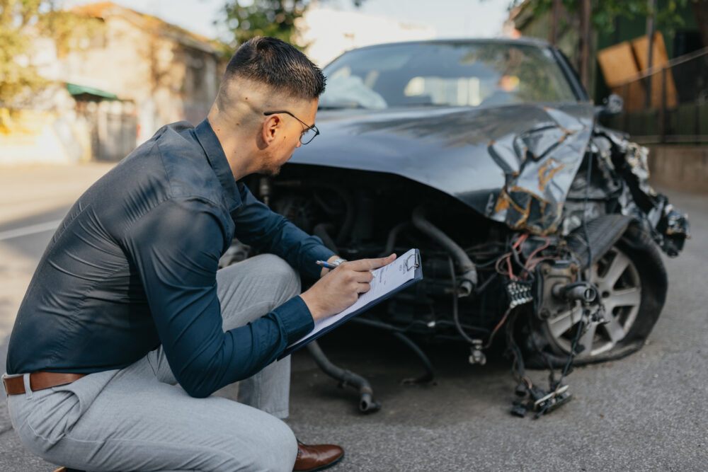 An insurance adjuster kneeling next to a damaged black car with a pen and clipboard.
