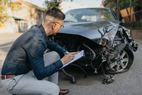 An insurance adjuster kneeling next to a damaged black car with a pen and clipboard.