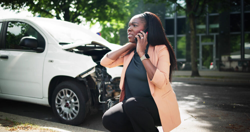 A distressed woman on the phone near her damaged white car after an accident.
