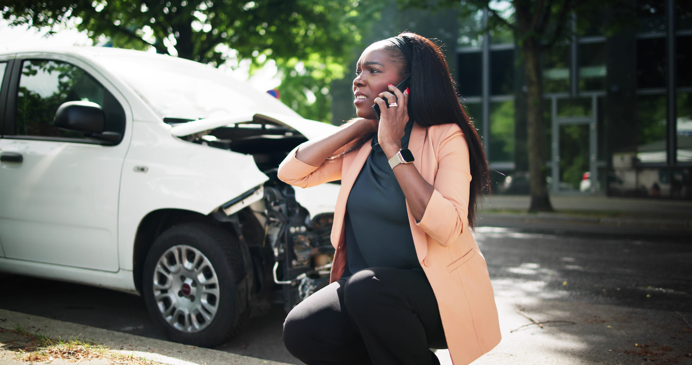 A distressed woman on the phone near her damaged white car after an accident.