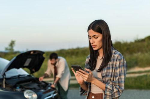A woman uses her phone while a man looks under the hood after a car accident.